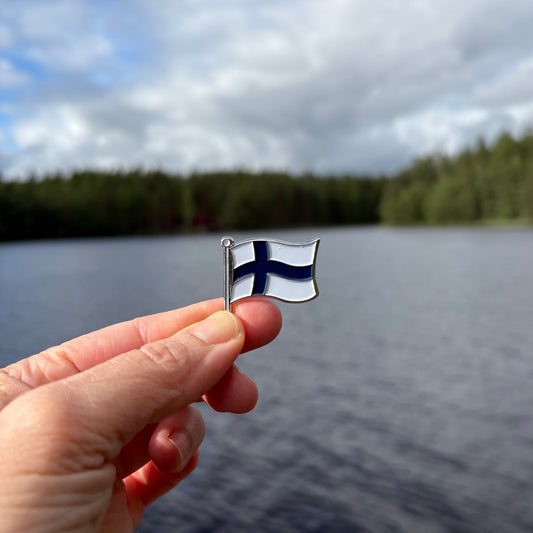 A hand holding a Finnish Flag enamel pin in front of a lake and forest