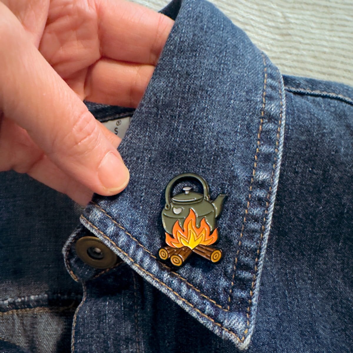 Campfire pot enamel pin clipped to a denim jacket lapel