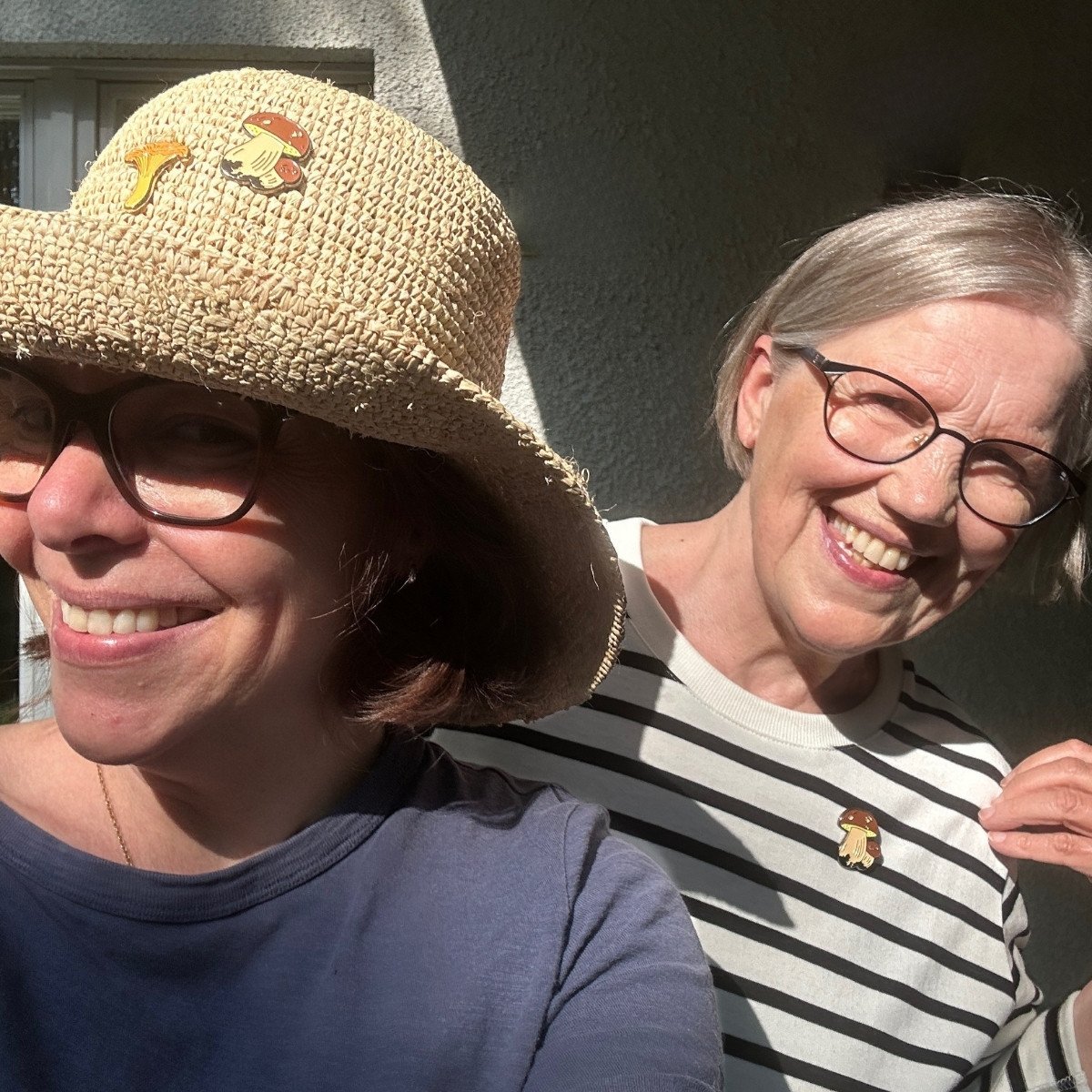 Two women wearing Kantarelli enamel pins. | Kaksi naista olkihatuissa, joissa Kantarelli-pinssit.