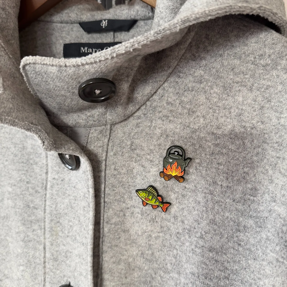 Close-up of perch and campfire pot enamel pins on a grey coat