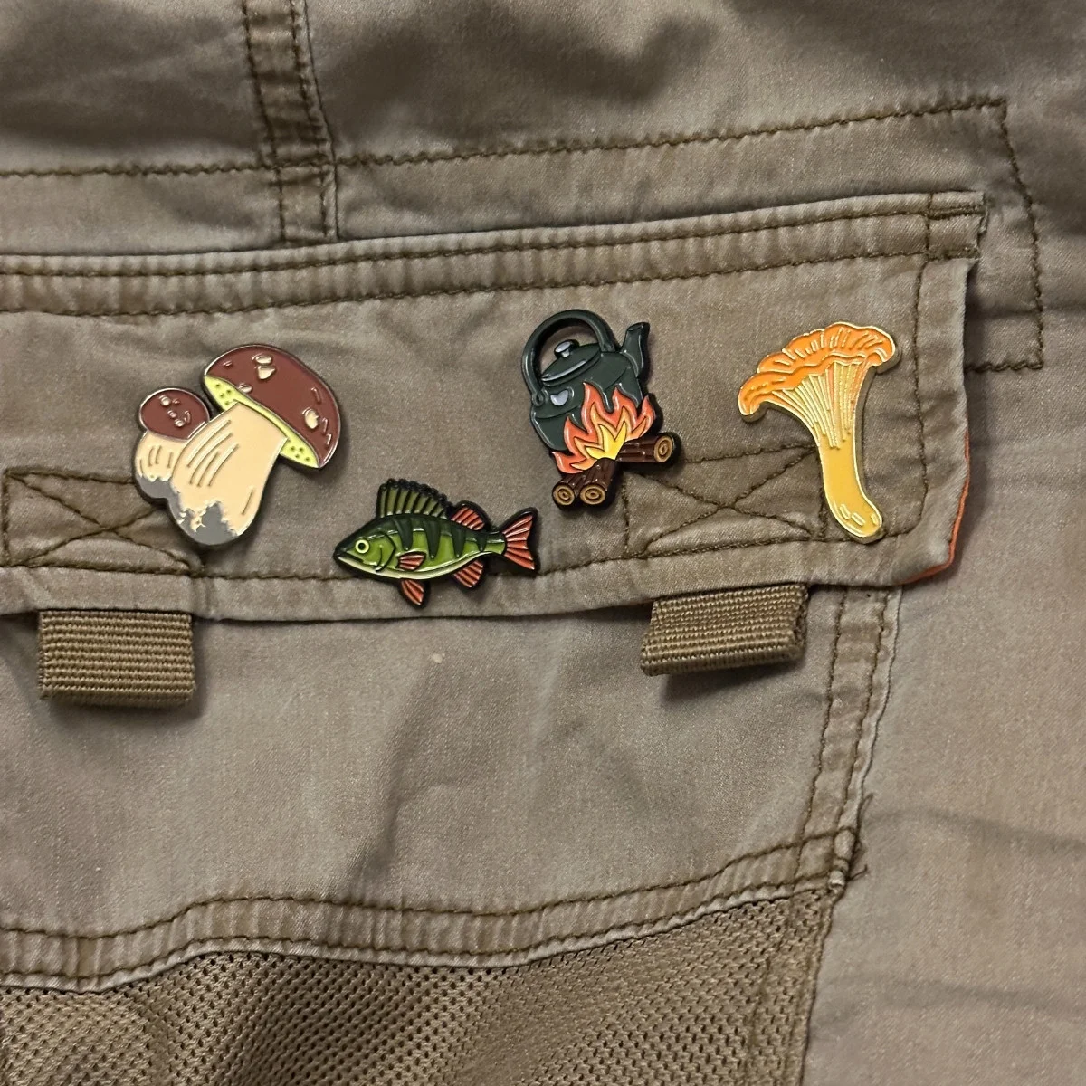 Set of Tarinapala enamel pins (Herkkutatti, Ahven, Nokipannu, Kantarelli) on jacket pocket.